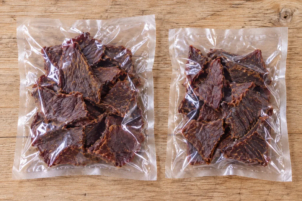 Две прозрачные закрытые упаковки с сушёными мясными ломтиками на деревянном столе